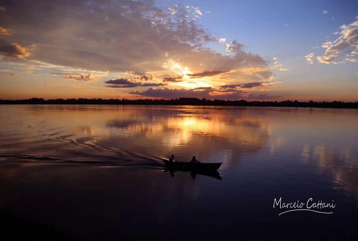 A small boat crossing the Uruguay River at first light, clouds reflected in still water