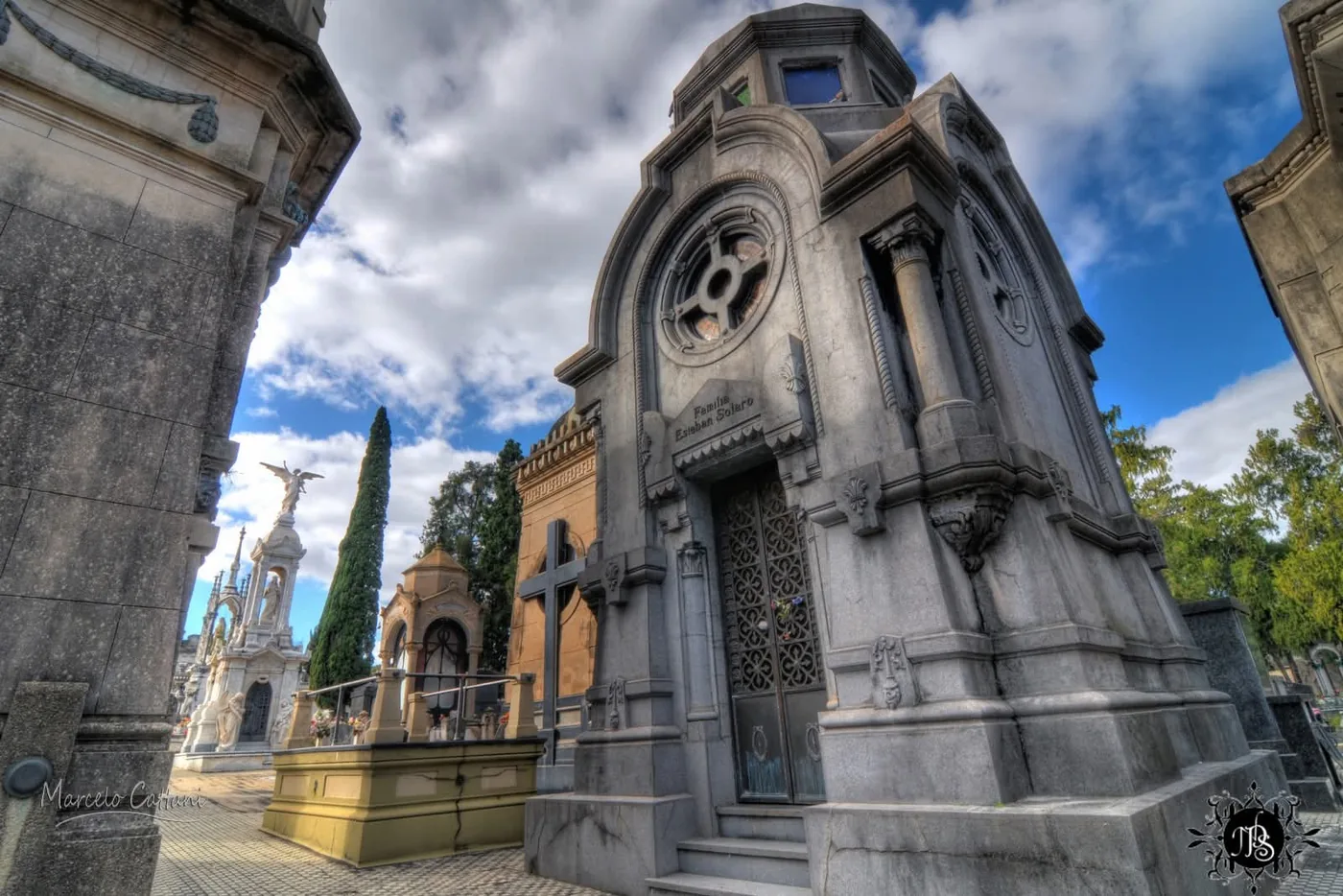 A granite family mausoleum with an iron cross, seen from below under a cloudy sky, Central Cemetery of Salto, Uruguay